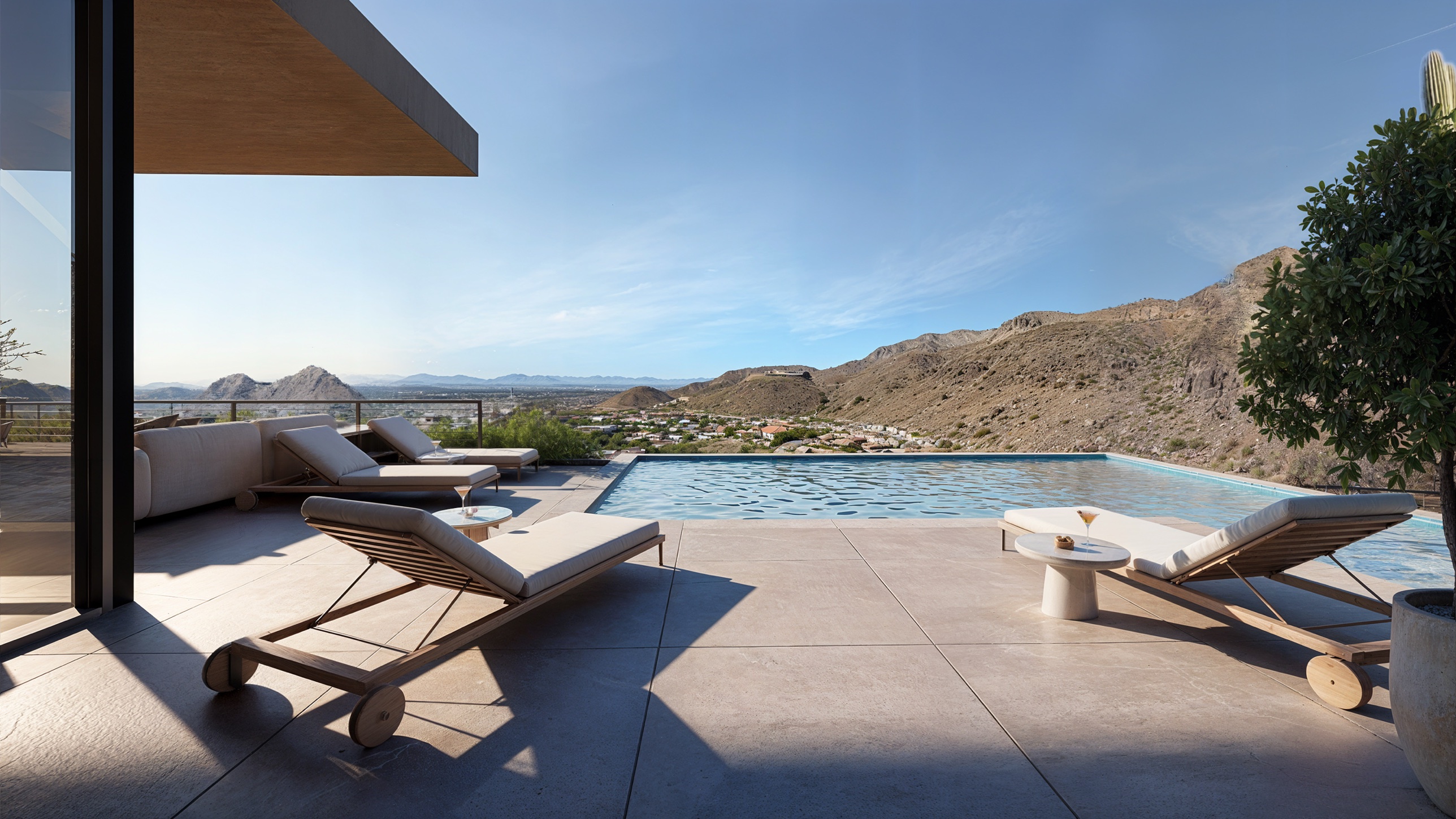 Wide view of the pool deck — pale stone paving to the pool edge, teak chaise lounges and side tables, the hillside and valley floor beyond the glass rail